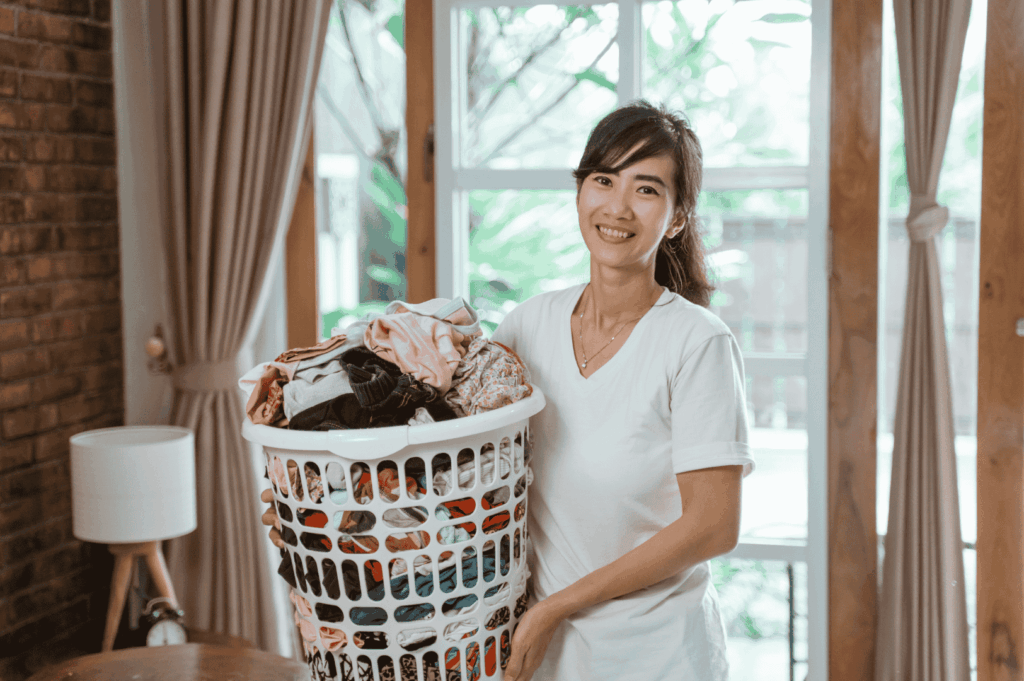 Smiling person holding a laundry basket indoors, representing domestic help in Singapore; ideal for expats navigating the hiring process of a domestic helper or foreign domestic worker through a maid agency or employment agency. Highlights key considerations like work permit application, helper's salary, transfer maids, levy concession, hiring one helper or subsequent FDW, and understanding MOM regulations. Depicts household responsibilities, family support, and the role of a housekeeper in taking care of children and relatives. Useful for employers deciding to hire, manage costs, pay, fees, and insurance, while ensuring fair employment, avoiding illegal candidates, and accessing services with expertise. Emphasizes the importance of being prepared, communicating obligations, and connecting with agencies to recover income, extend control, and save money. A complete guide to domestic worker jobs, manpower demand, and penalties involved in Singapore.