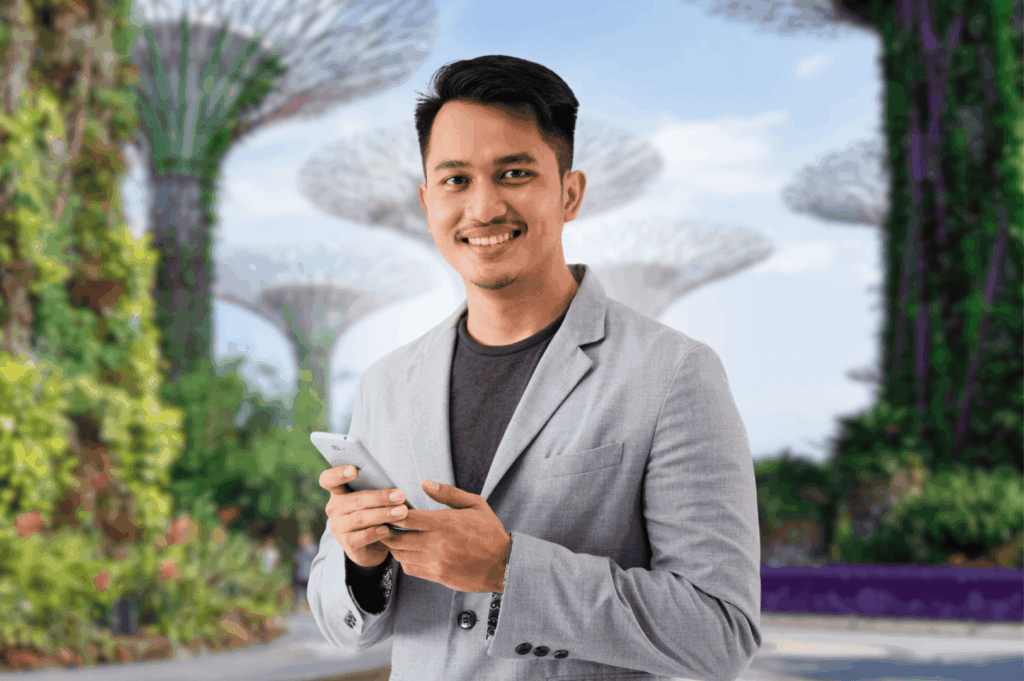 Smiling person holding a smartphone in front of Singapore’s iconic Supertree Grove at Gardens by the Bay, symbolizing the ease of exploring Singapore with its mass rapid transit system. This Singapore MRT Guide for Expats highlights MRT rides across MRT lines including the North South Line, East West Line, Circle Line, Downtown Line, North East Line, Thomson East Coast Line, and LRT lines like Bukit Panjang LRT. Expats can access MRT stations and train stations such as Dhoby Ghaut, City Hall, Chinatown MRT, Marina Bay, Orchard Road, Raffles Place, Botanic Gardens, and Tanjong Pagar during peak hours, off peak hours, rush hours, off peak times, and midnight daily. Public transport options include public buses, bus rides, and train services with train frequency details and operating hours. Riders can pay directly and simply tap using contactless bank cards, mobile wallets like Apple Pay, Samsung Pay, Fitbit Pay, or physical cards such as EZ-Link, NETS FlashPay, and travel cards from other countries. Ticketing machines offer single trip tickets and the Singapore Tourist Pass. The MRT map supports cost-effective journey planning with Google Maps, SimplyGo app, MyTransport app, and the official app. Features include priority seats, admin fee info, first bus timings, convenience stores, and interchange stations for easy access across one line or total distance traveled.