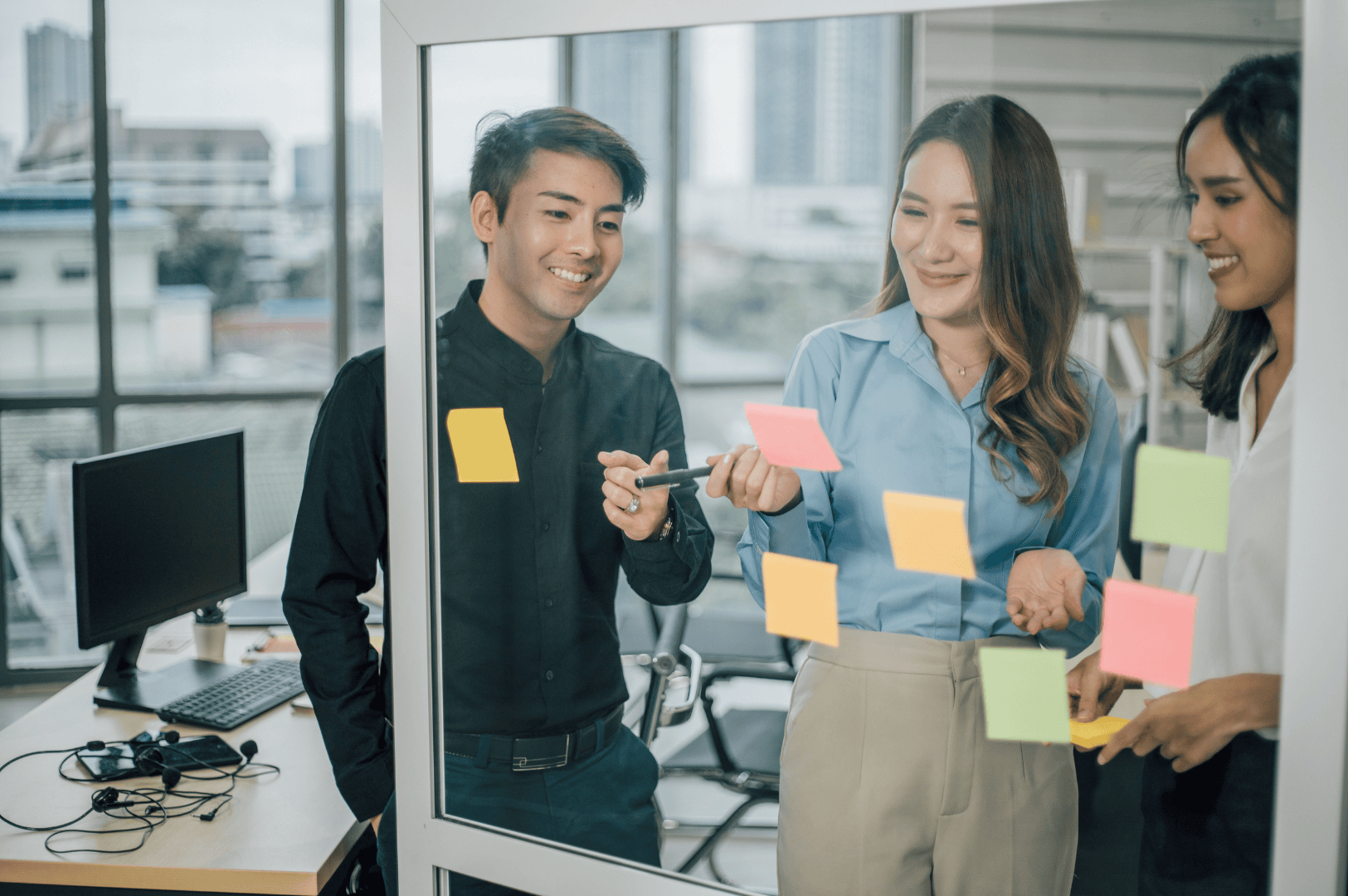Three professionals gather around a glass wall covered in colorful sticky notes, immersed in a collaborative brainstorming session—capturing Singapore’s emphasis on collective problem-solving, visual planning, and respectful dialogue. The open office setting and city views reflect transparency, modernity, and the unspoken rhythm of urban work life.