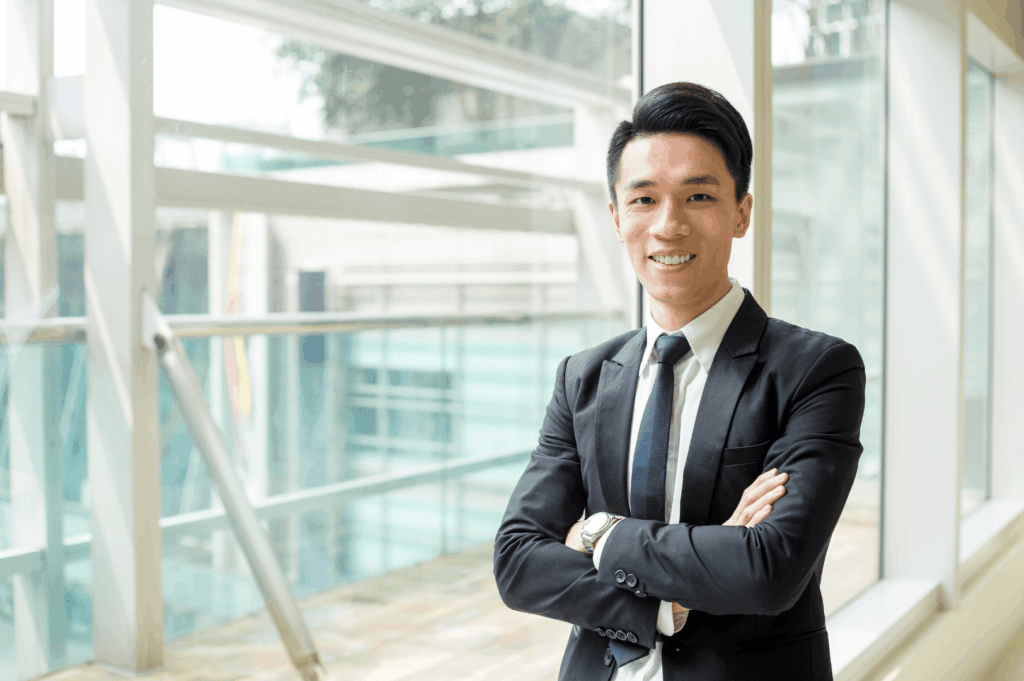 A poised professional stands confidently in a sunlit modern building, arms crossed and smiling—embodying Singapore’s polished work culture, where formality meets approachability. The architectural backdrop and greenery reflect the city’s fusion of precision, transparency, and biophilic design in corporate environments.