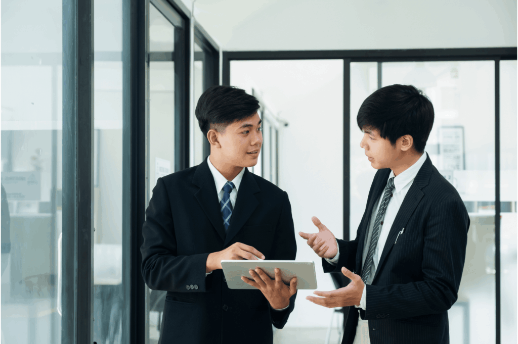 Two professionals in business attire engage in a focused discussion in a sleek office setting—one presenting via tablet, the other responding with expressive gestures. The scene reflects Singapore’s tech-forward communication style, hierarchical nuance, and the subtle choreography of respect and responsiveness in workplace interactions.