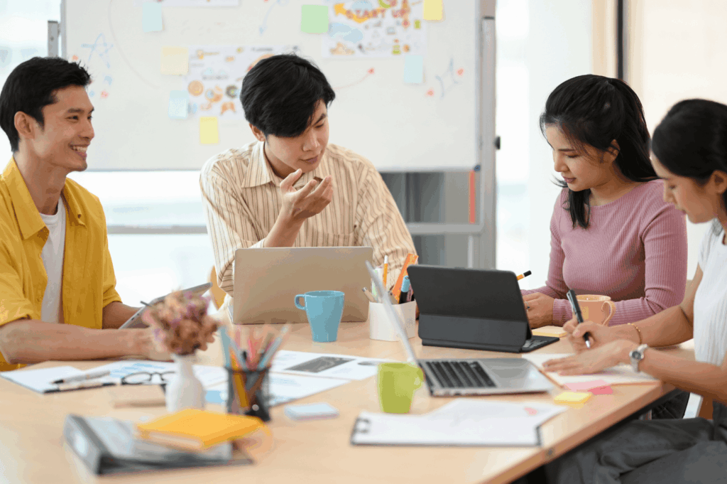 Four colleagues seated around a shared table, immersed in laptops, notebooks, and casual conversation—capturing Singapore’s balance of productivity and comfort in collaborative spaces. Coffee mugs, stationery, and a flower vase soften the scene, while the whiteboard behind them hints at structured planning and the quiet rigor of team-based problem-solving.