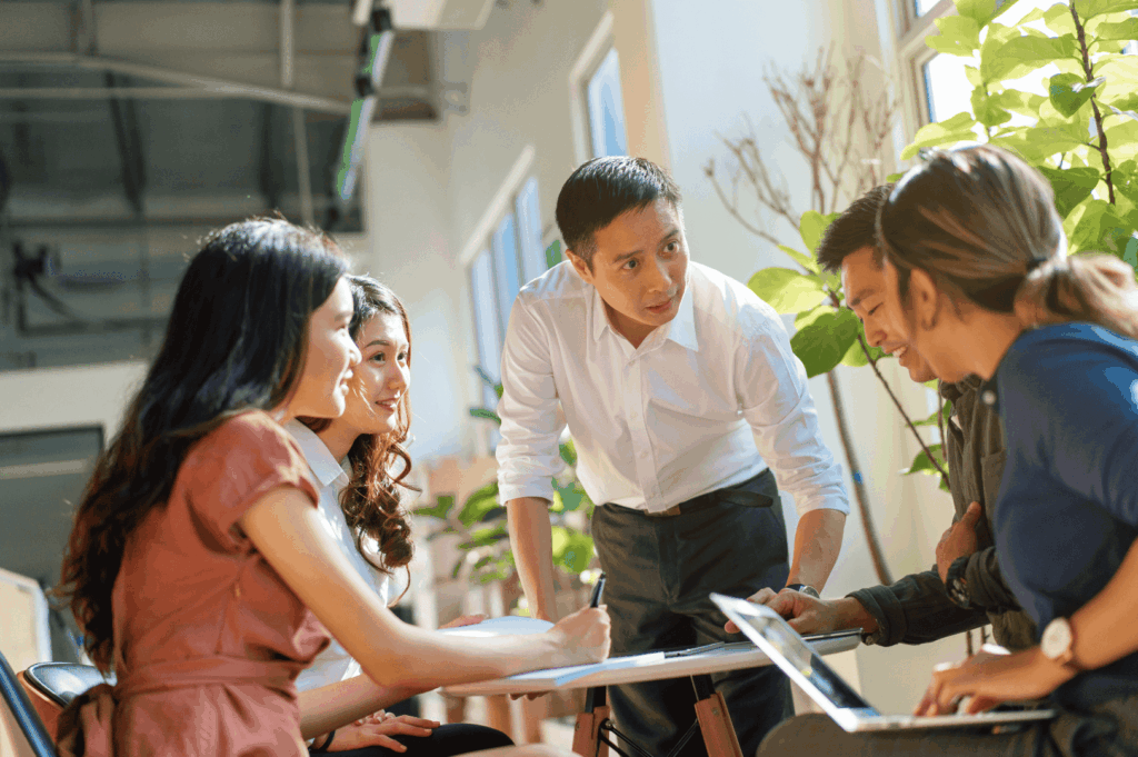 A team of five professionals engaged in a dynamic meeting, with one standing to lead the discussion while others listen, take notes, and work on laptops—illustrating Singapore’s respect for structured leadership, attentive collaboration, and the subtle hierarchy of group dynamics. Natural light and greenery soften the scene, reflecting the city’s growing embrace of wellness and biophilic design in modern workspaces.