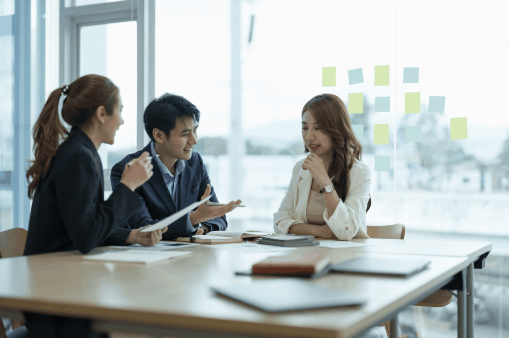 Three professionals seated around a table in a bright office, engaged in thoughtful discussion with gestures, notes, and attentive listening—reflecting Singapore’s culture of respectful dialogue, strategic planning, and the quiet choreography of team dynamics. Sticky notes on the glass wall behind them hint at structured ideation and visual organization.