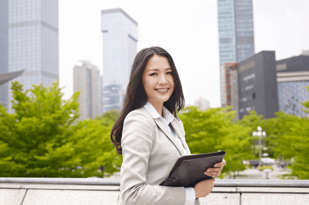 A confident professional stands outdoors in a vibrant cityscape, holding a folder and smiling—embodying Singapore’s fusion of ambition, approachability, and urban sophistication. Surrounded by greenery and high-rise architecture, the scene reflects the balance between nature, career drive, and the polished public persona often expected in the city’s work culture.