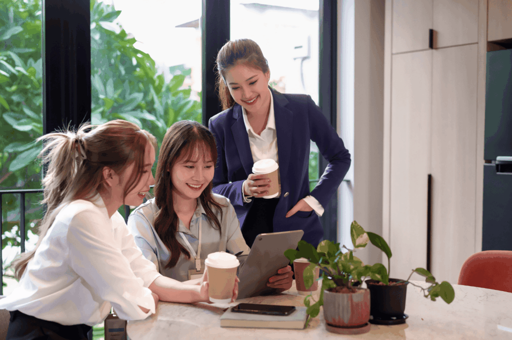 hree professionals gathered around a table, sharing coffee and reviewing a tablet—capturing Singapore’s blend of informal meetings, tech-savvy collaboration, and the understated rituals that foster trust and productivity. The bright setting, greenery, and smart-casual attire reflect a modern work culture that values both comfort and clarity.