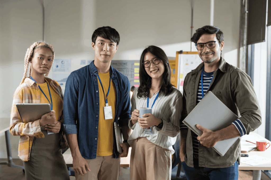 Four team members stand in a collaborative workspace, each holding tools of their trade—tablet, notebook, coffee mug, and folder—symbolizing Singapore’s multifaceted work culture rooted in preparedness, diversity, and shared ownership. Lanyards and whiteboards filled with sticky notes evoke a structured yet dynamic environment where innovation thrives through respectful teamwork.