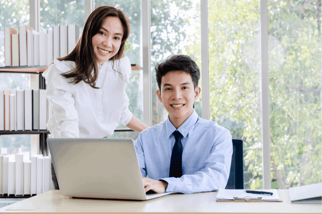 Two professionals in a bright office setting share a moment of collaboration—one seated at a desk working on a laptop, the other standing nearby with a warm smile. Surrounded by greenery and organized shelves, the scene reflects Singapore’s emphasis on teamwork, professionalism, and the subtle social cues that shape everyday workplace interactions.