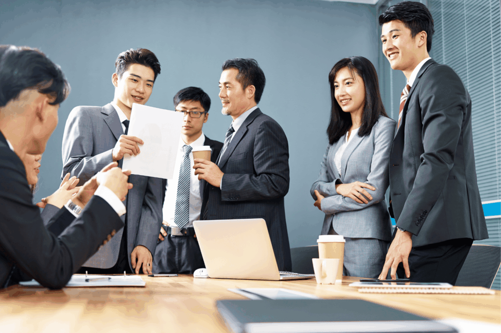 Eight professionals gather around a conference table in a modern office, exchanging ideas over coffee, laptops, and documents—capturing Singapore’s culture of structured collaboration, clear communication, and the quiet choreography of group dynamics. The mix of seated and standing participants reflects fluid leadership roles and the respectful engagement expected in formal business settings.