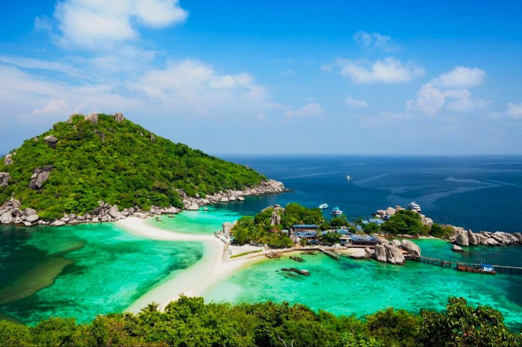 Scenic view of a tropical island with lush greenery, turquoise waters, and a sandy beach under a bright blue sky. Boats are visible in the distance.