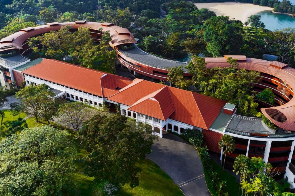 Aerial view of a large building with red roofs, surrounded by lush greenery and trees. A beach is visible in the background, creating a serene atmosphere.