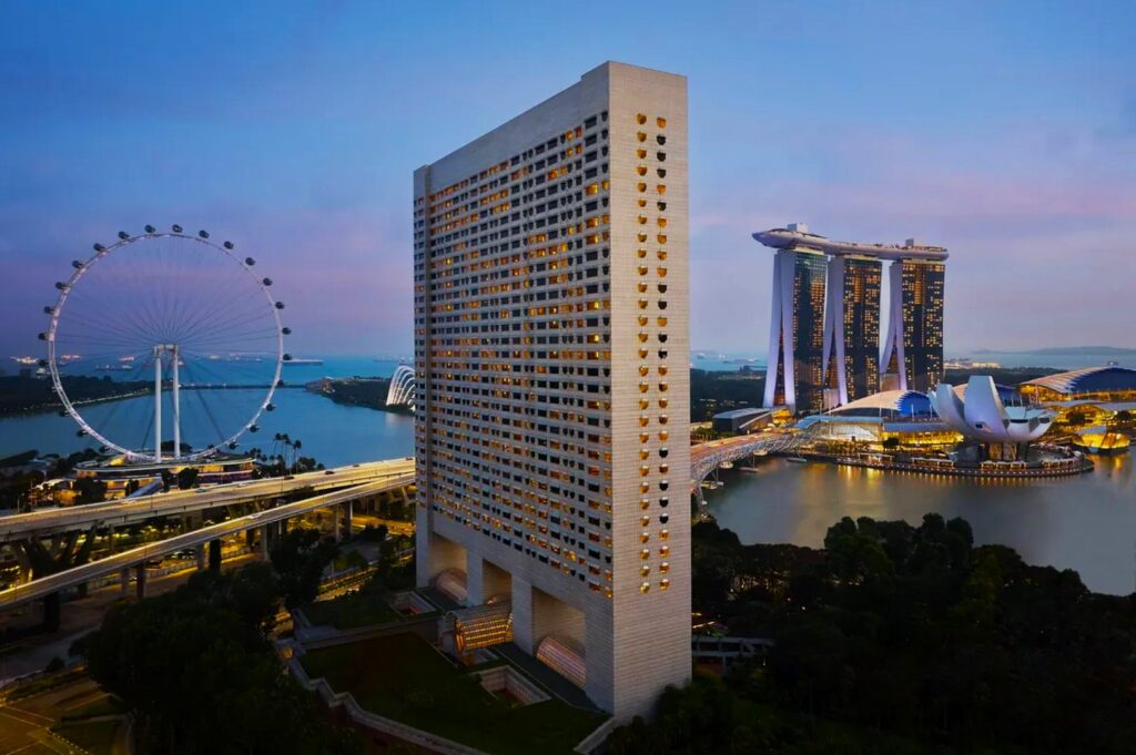 Cityscape at dusk with a tall, white high-rise building in the foreground. A ferris wheel and Marina Bay Sands are seen against a pastel sky.