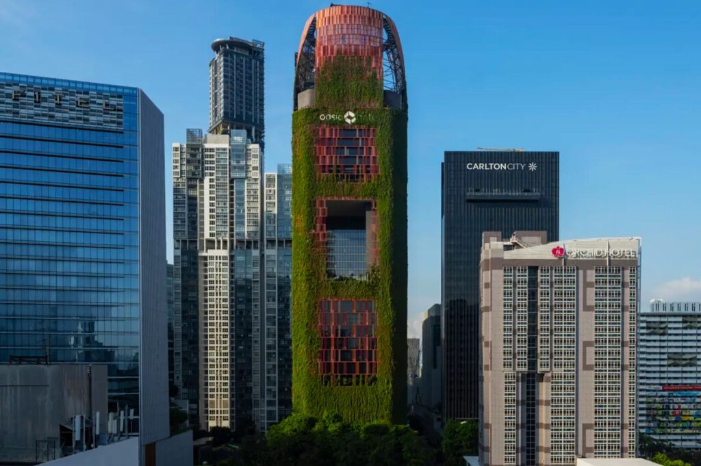 A tall building adorned with lush green vertical gardens and reddish-brown accents stands amidst modern skyscrapers under a clear blue sky.