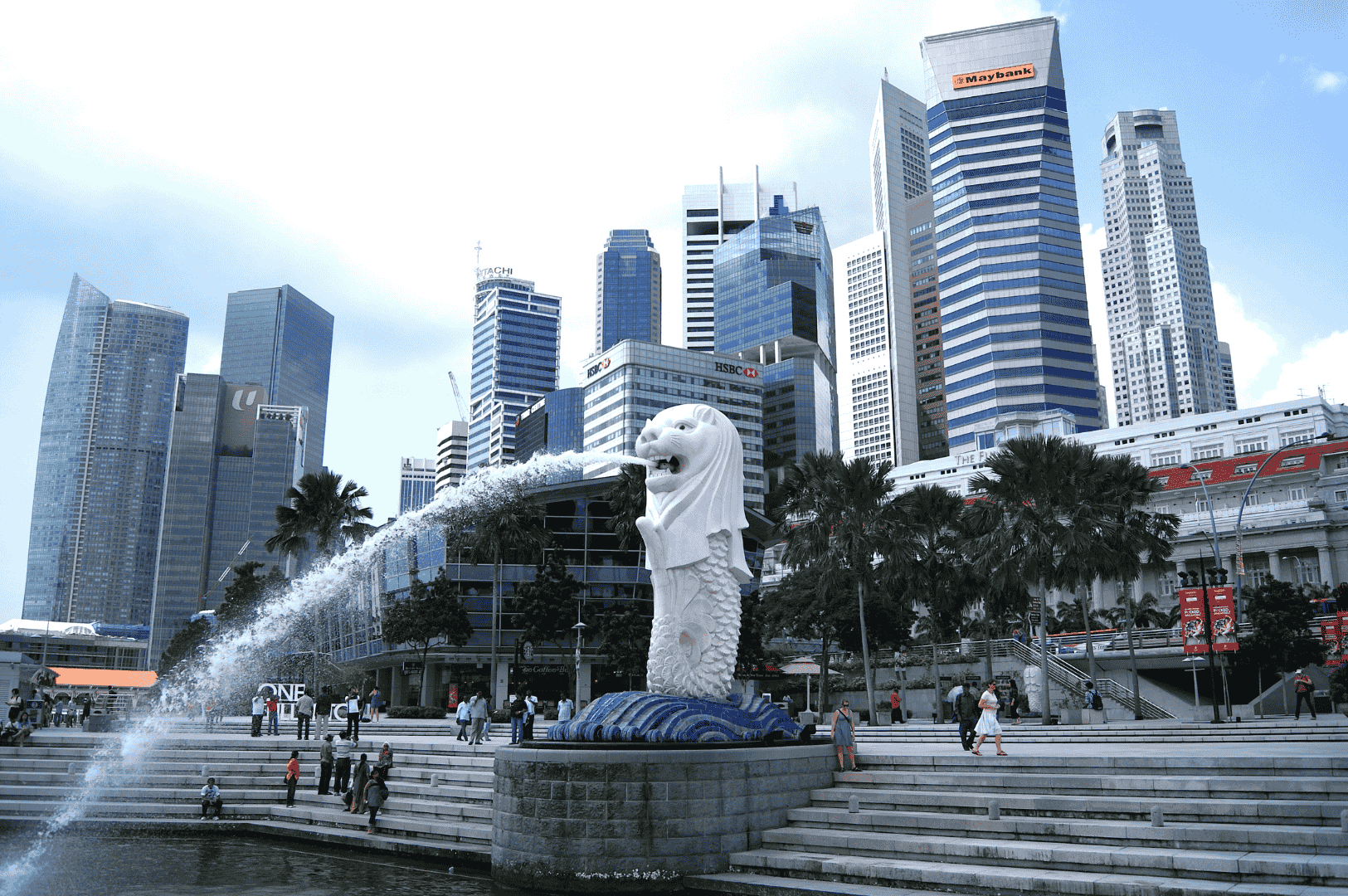 The Merlion statue spouts water into Marina Bay under a bright sky, framed by Singapore’s sleek financial skyline. Locals and tourists gather on the steps nearby, dressed in breathable, sun-ready outfits—light fabrics, short sleeves, and practical footwear—embodying the balance of comfort and style essential for navigating Singapore’s humid climate as an expat.