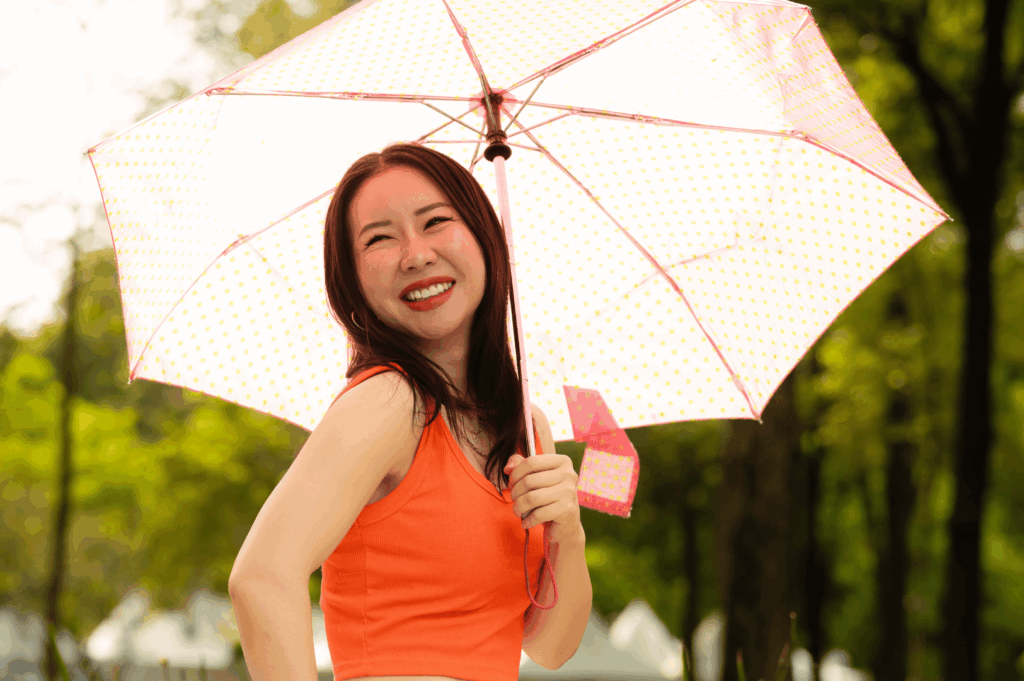 A smiling person stands outdoors under a transparent pink polka-dot umbrella, wearing a sleeveless orange top suited for Singapore’s humid climate. The lush greenery and bright sunlight evoke a cheerful, tropical vibe, while the outfit and umbrella reflect a practical yet expressive approach to dressing for sudden rain and intense heat—key considerations for expats building a versatile wardrobe.
