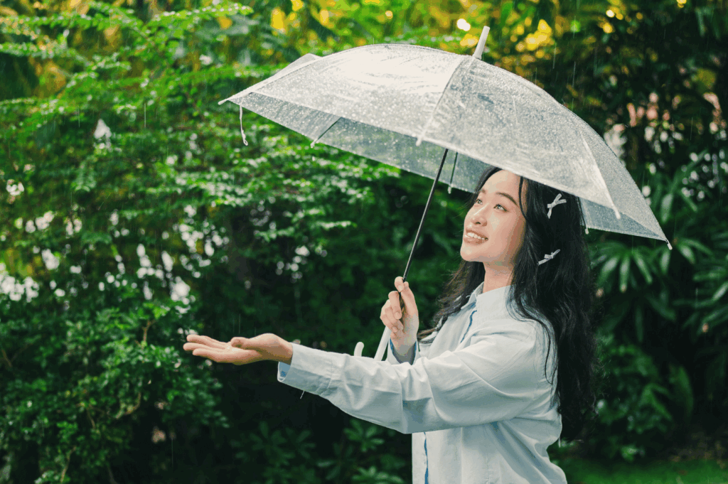 A smiling person stands in a lush garden under a transparent umbrella, wearing a light long-sleeved shirt and butterfly-shaped hair clips. Their outstretched hand and joyful expression capture a moment of playful connection with Singapore’s tropical rain. The outfit reflects a thoughtful expat wardrobe—light layers for humidity, rain protection, and personal flair that balances comfort with individuality