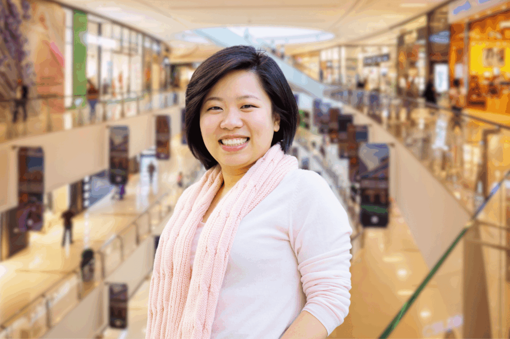 A smiling person stands in a brightly lit Singapore shopping mall, wearing a light pink top and matching scarf on skin—an airy, coordinated outfit suited for indoor-outdoor transitions in the city’s humid climate. The modern architecture and bustling atmosphere highlight the need for breathable fabrics and adaptable layers in an expat wardrobe built for comfort, style, and spontaneity.