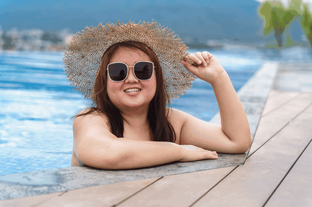 A person relaxes at the edge of a modern pool, smiling beneath oversized sunglasses while holding a wide-brimmed straw hat. The tropical backdrop of palm trees and mountains evokes resort-style serenity. The swimwear and accessories reflect a chic yet functional expat wardrobe—sun-shielding, breathable, and perfect for embracing Singapore’s heat with effortless elegance.