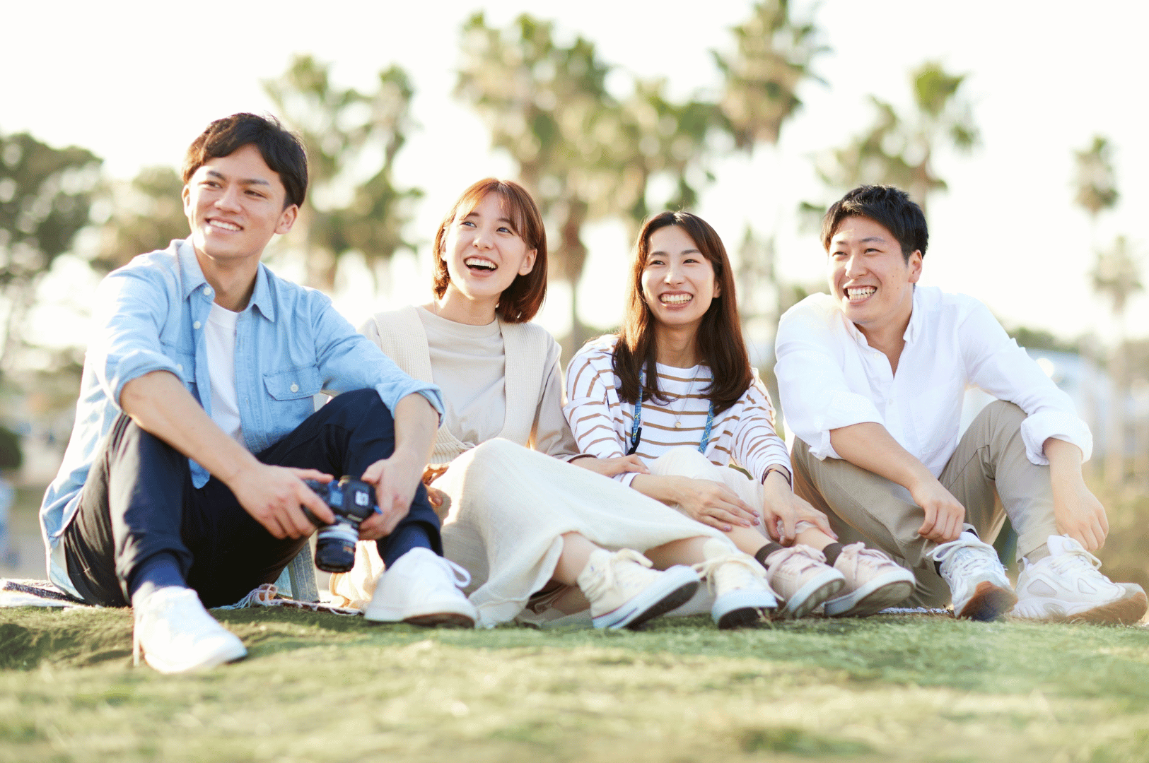 Four friends sit together on a grassy patch beneath palm trees, smiling and relaxed in the tropical daylight. One holds a camera, suggesting a shared moment of documentation or storytelling. Their casual attire and natural ease reflect the informal warmth and communal spirit often found in Southeast Asian gatherings. The open-air setting and joyful expressions evoke the region’s emphasis on connection, hospitality, and the value of spending time outdoors with loved ones.