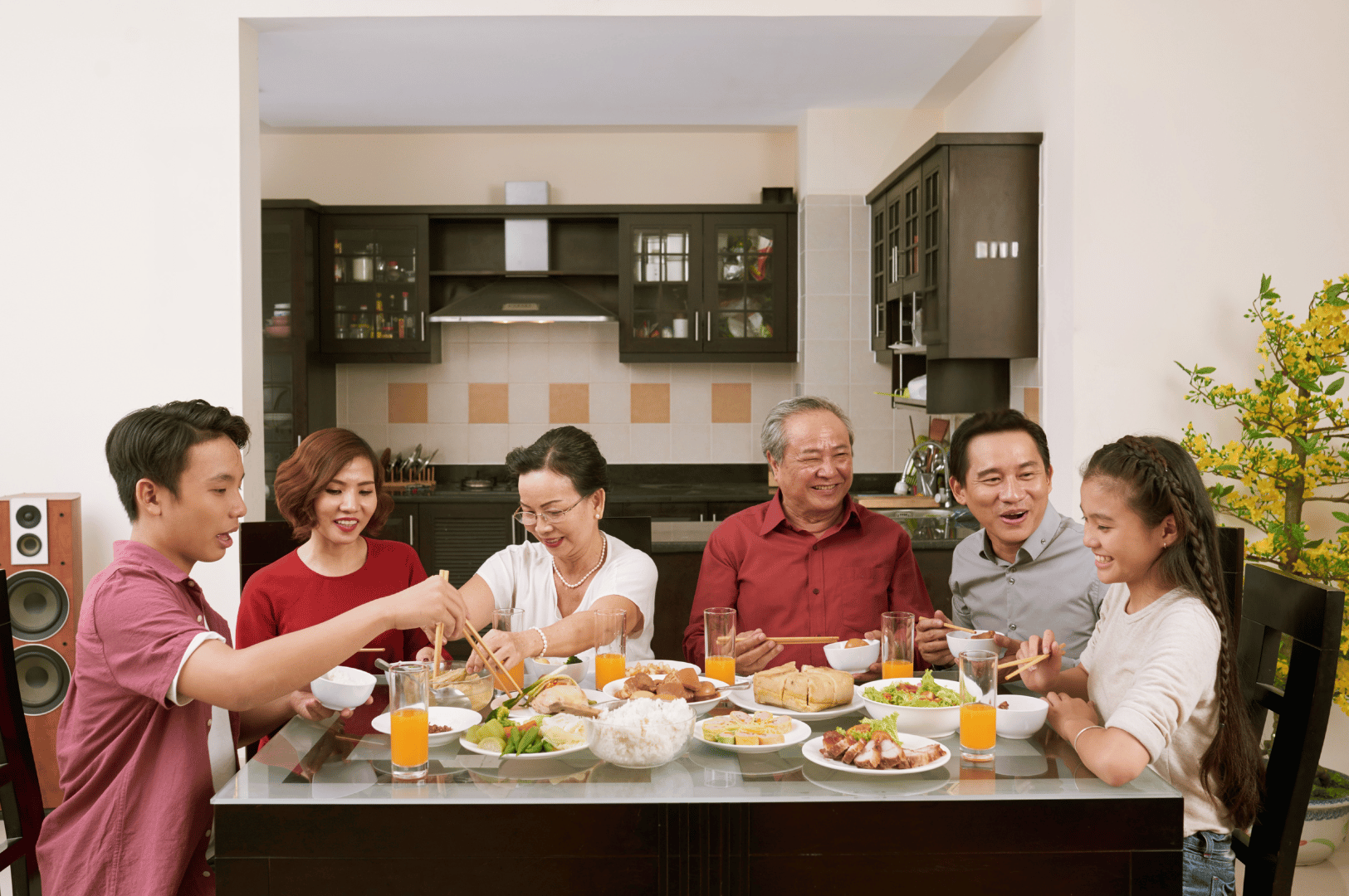 Six people gather around a dining table in a modern home, sharing a meal of rice, vegetables, and meat with glasses of juice and water. Their engaged expressions and communal gestures reflect the cultural importance of shared meals in Southeast Asia, where hospitality and togetherness are central to social etiquette. The warm lighting and casual setting evoke a sense of familial intimacy, highlighting how food rituals often serve as a bridge for expats to connect with local traditions.