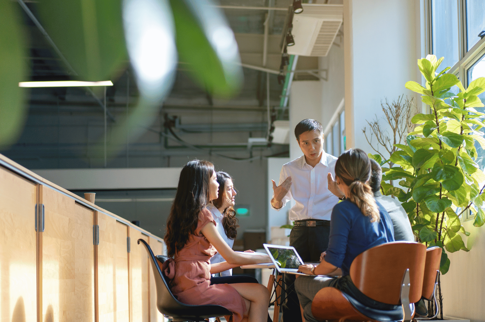 Five colleagues engage in a lively discussion in a sunlit modern office, with one person standing and gesturing while others listen attentively around a table. A laptop and green plants add context to the collaborative, tech-savvy environment. This scene reflects Southeast Asia’s evolving professional etiquette, where respectful listening, expressive communication, and group consensus are valued in both local and expat work cultures.