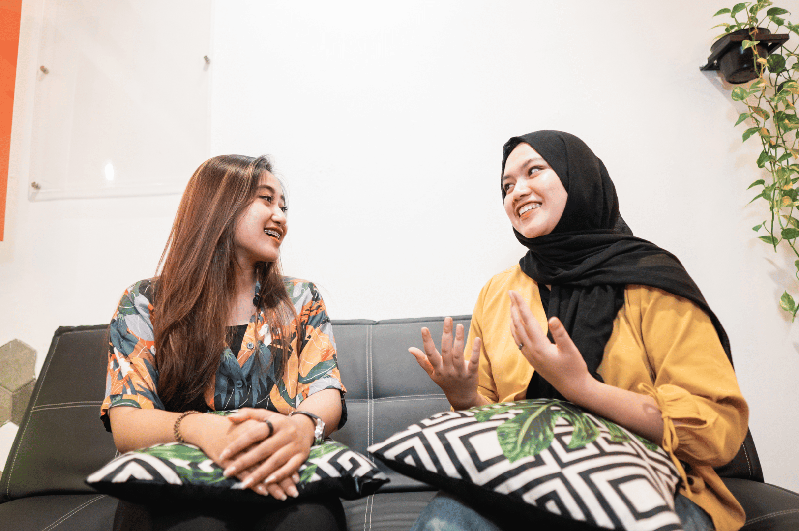 Two individuals sit on a dark couch in a casual indoor setting, engaged in animated conversation. One wears a floral shirt, the other a black hijab and mustard yellow top—both holding leaf-patterned pillows. Their expressive gestures and attentive posture reflect the region’s emphasis on respectful dialogue, active listening, and interpersonal warmth. The minimalist decor and small wall-mounted plant add a touch of intimacy, highlighting how everyday spaces foster meaningful social exchange in Southeast Asian culture.