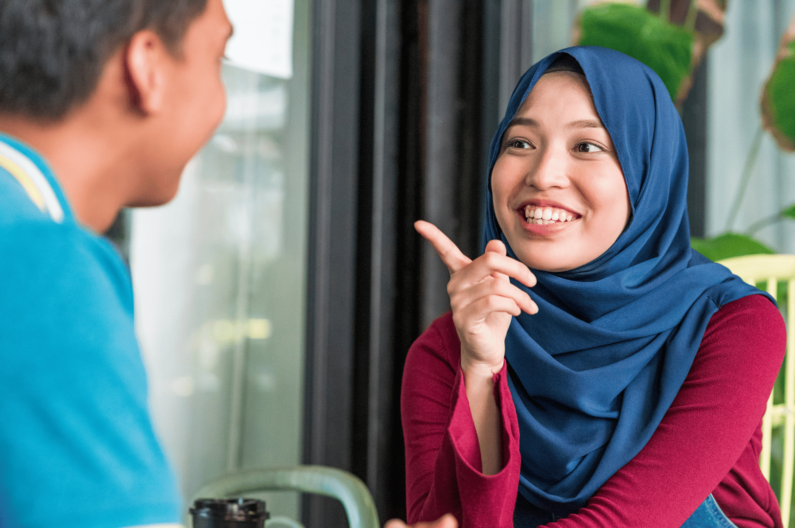 Smiling woman in blue hijab animatedly chatting with a friend in a cozy café setting, gesturing with playful confidence; a candid moment that captures the warmth and expressiveness of everyday Singaporean conversation—ideal for illustrating how Singlish brings personality and connection to local interactions.