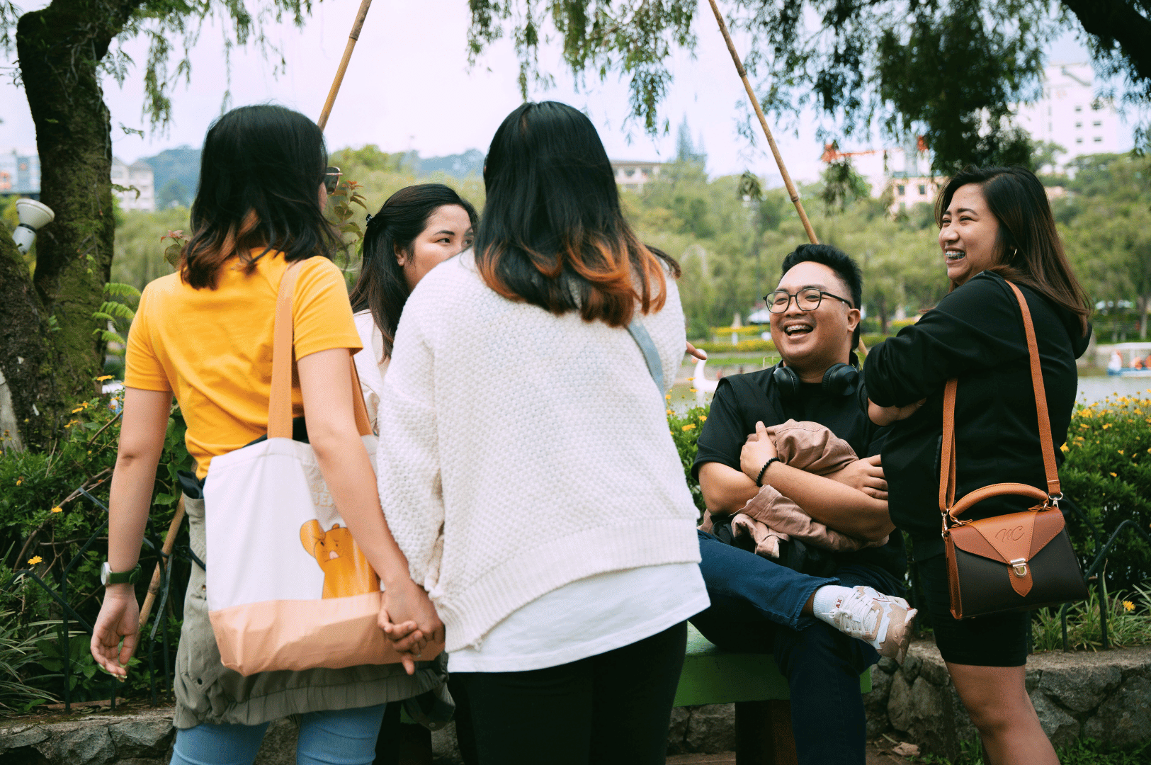 Group of young adults laughing and chatting in a lush urban park, with one person seated on a bench holding a jacket and smiling broadly; a lively, candid moment that captures the spirit of Singaporean camaraderie and the expressive charm of Singlish in everyday social banter.