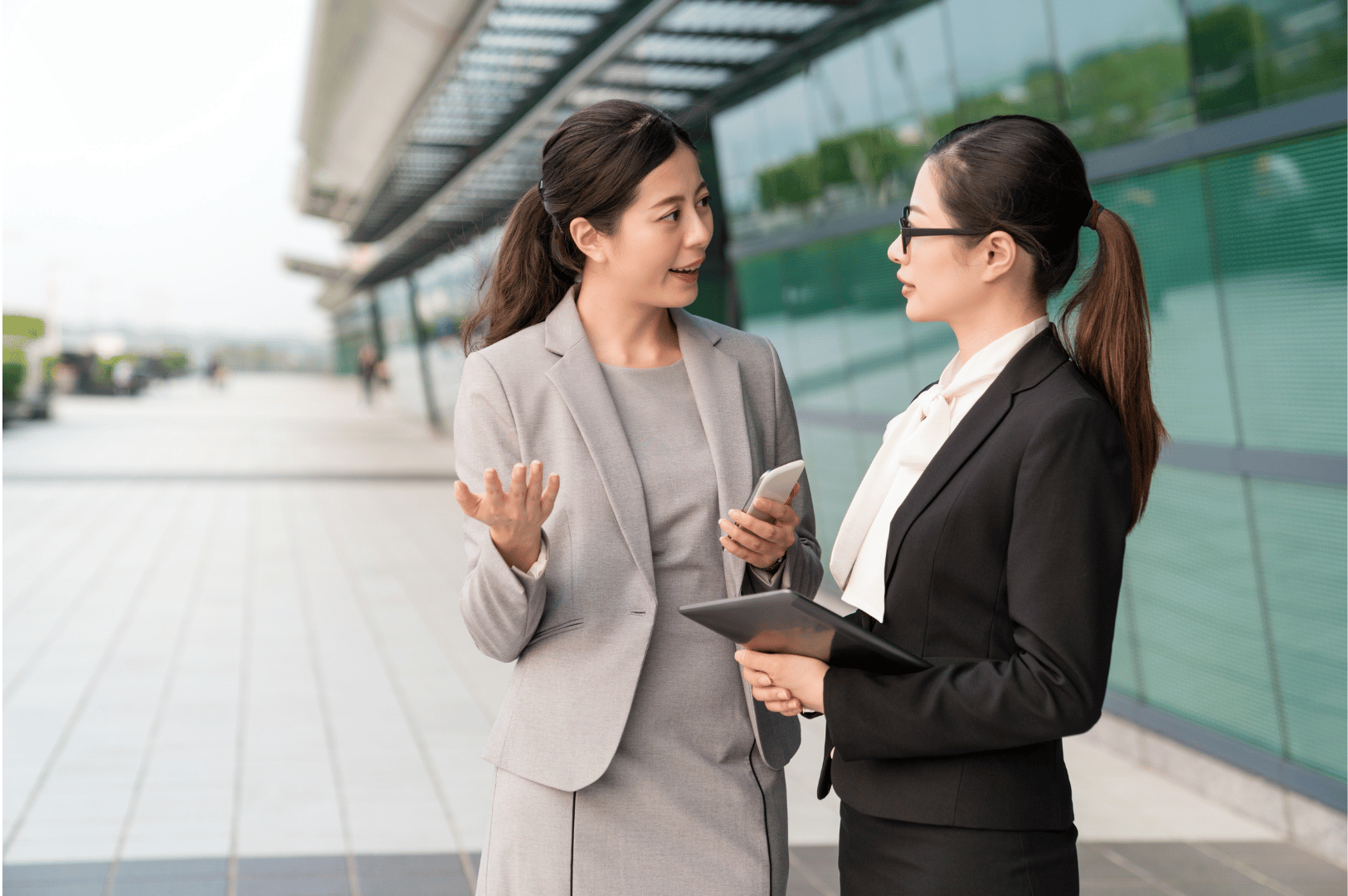 Two professionally dressed women conversing outside a sleek glass-fronted building, one holding a smartphone and the other a tablet; a polished, modern scene that reflects how Singlish phrases like “lah” and “can” seamlessly blend into Singapore’s business culture, adding local flavor to corporate communication.