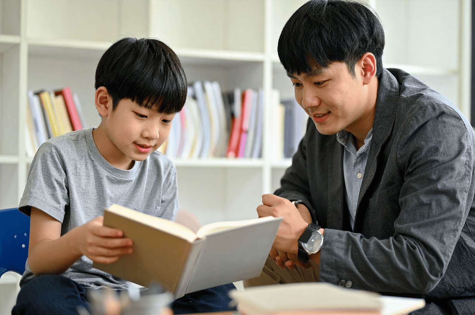 Young boy in a grey t-shirt reads aloud from a book beside an attentive adult mentor, seated in front of a bookshelf—capturing the heart of Singapore’s tuition culture through personalized guidance, intergenerational learning, and the emotional intimacy of one-on-one education.