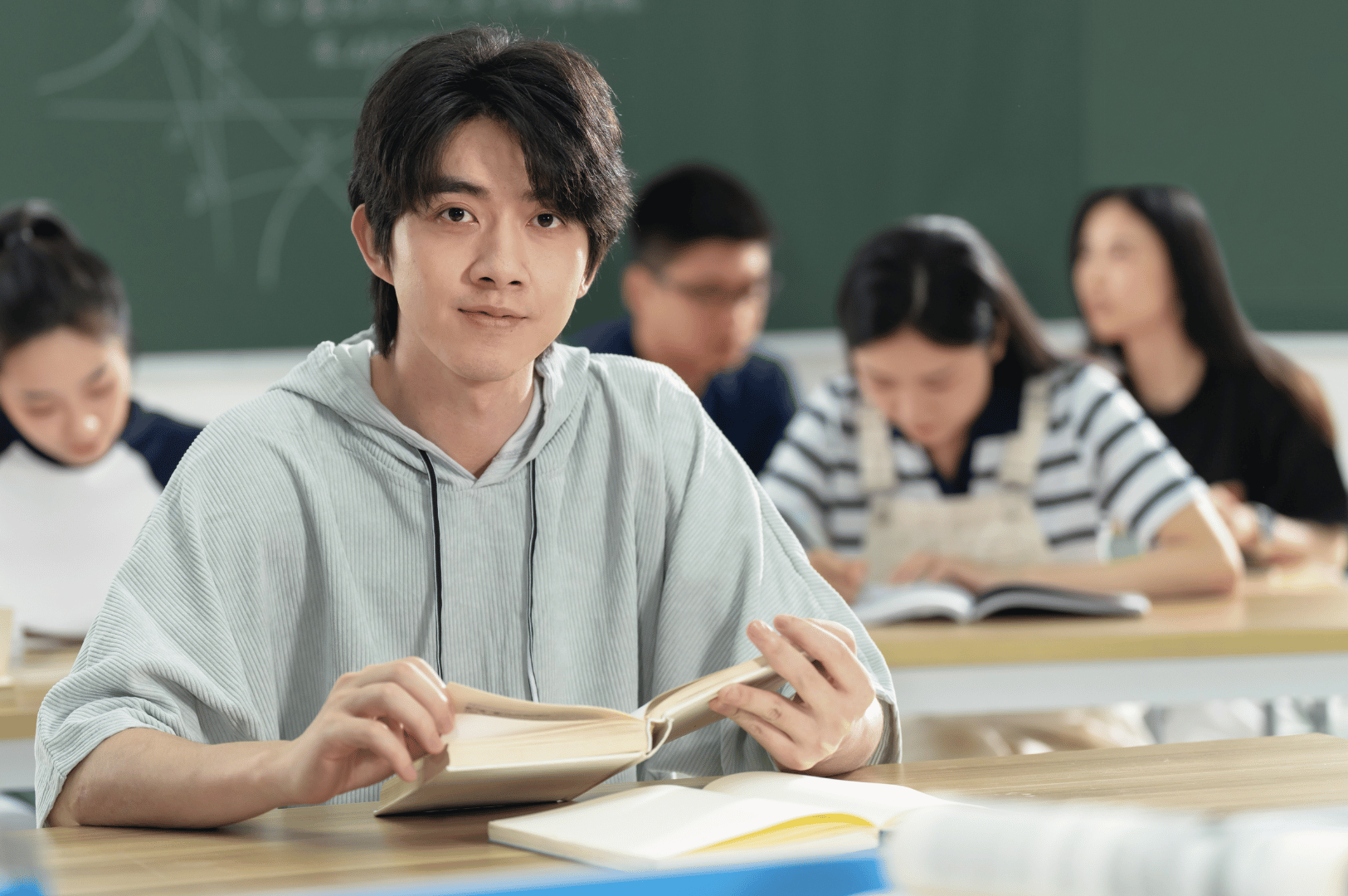 Student in light hoodie gazes directly at the camera while holding an open book in a focused classroom—framed by peers immersed in study and a chalkboard marked with geometry sketches. The image evokes the intensity and normalization of academic rigor in Singapore’s supplementary education landscape.