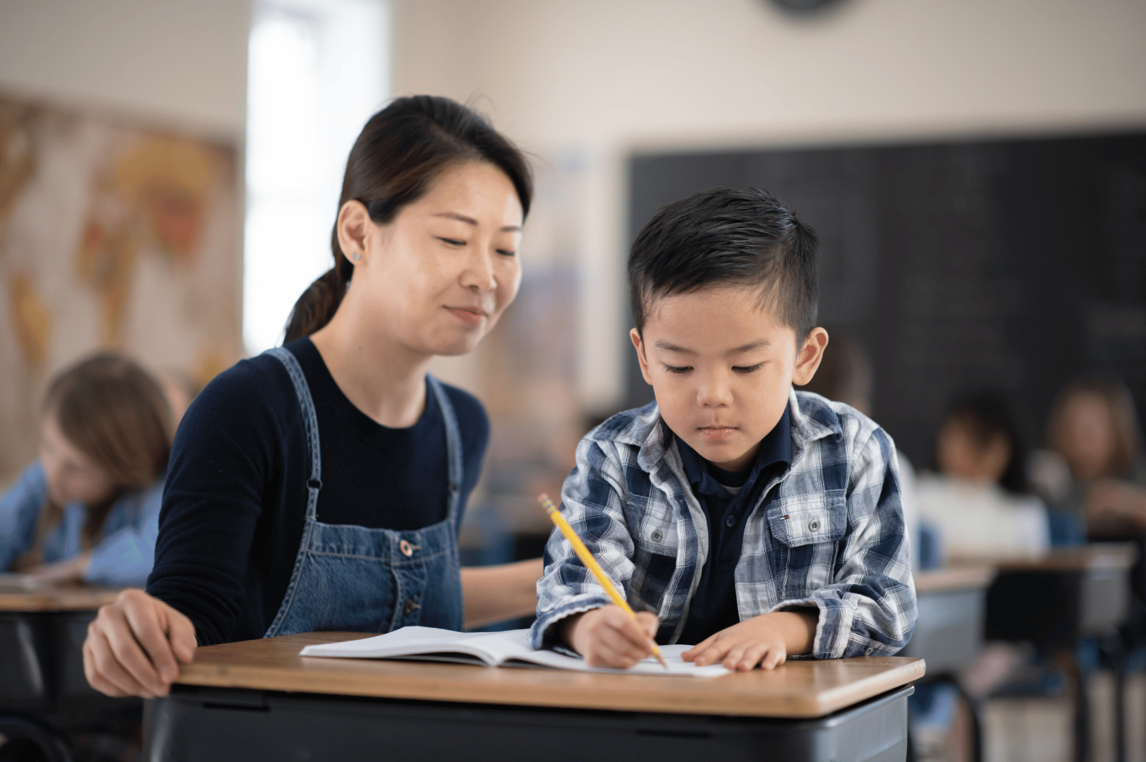 Young child in plaid shirt concentrates on writing in a notebook, guided by a gentle adult mentor in denim overalls—set in a warm, collaborative classroom. The image reflects the nurturing side of Singapore’s tuition culture, where early academic support and emotional connection shape foundational learning experiences.