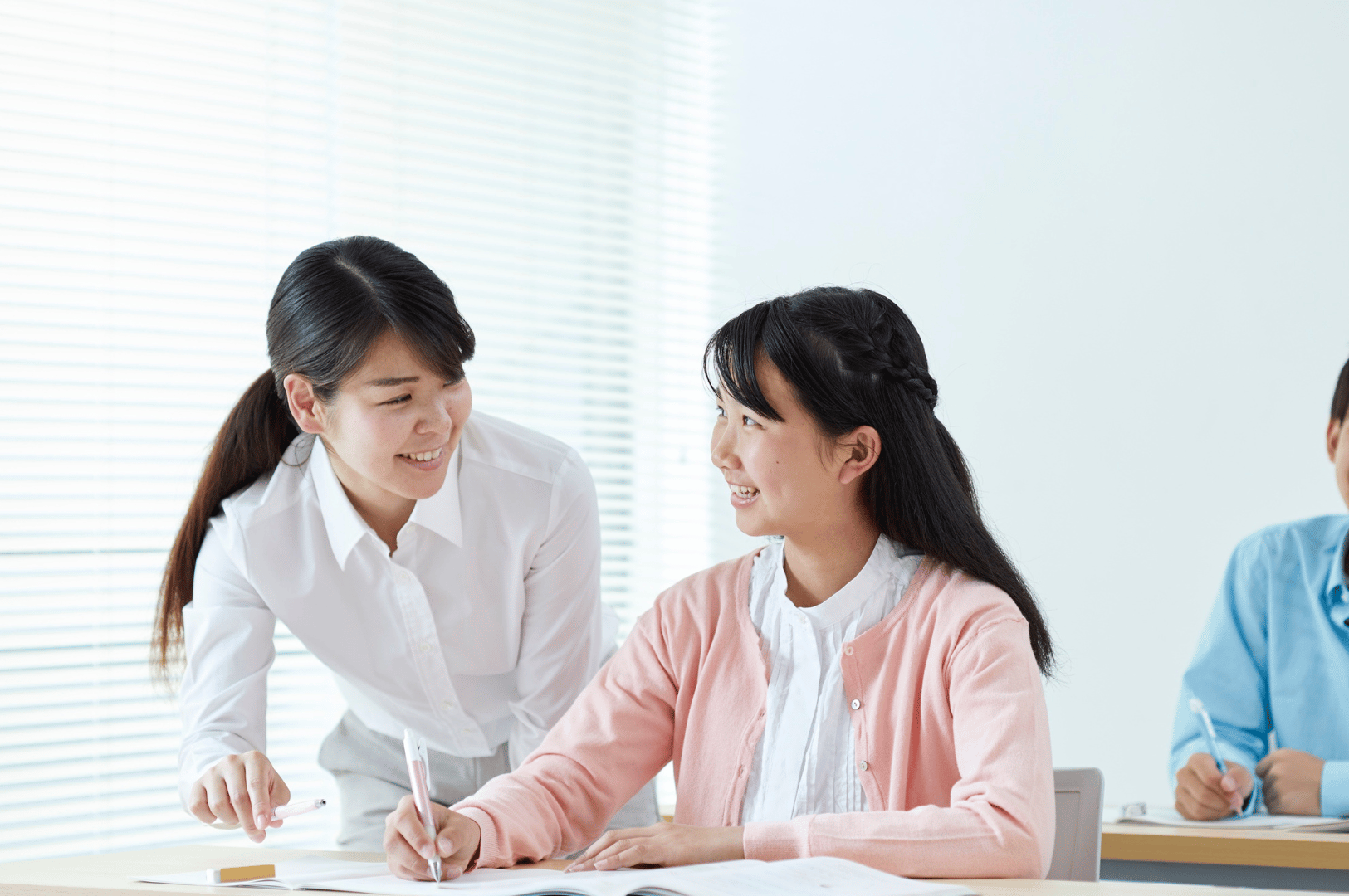 Smiling student in a light pink cardigan receives guidance from a supportive teacher in a sunlit classroom—capturing the relational warmth and personalized attention that define Singapore’s tuition culture, where academic success is nurtured through trust, encouragement, and one-on-one mentorship.