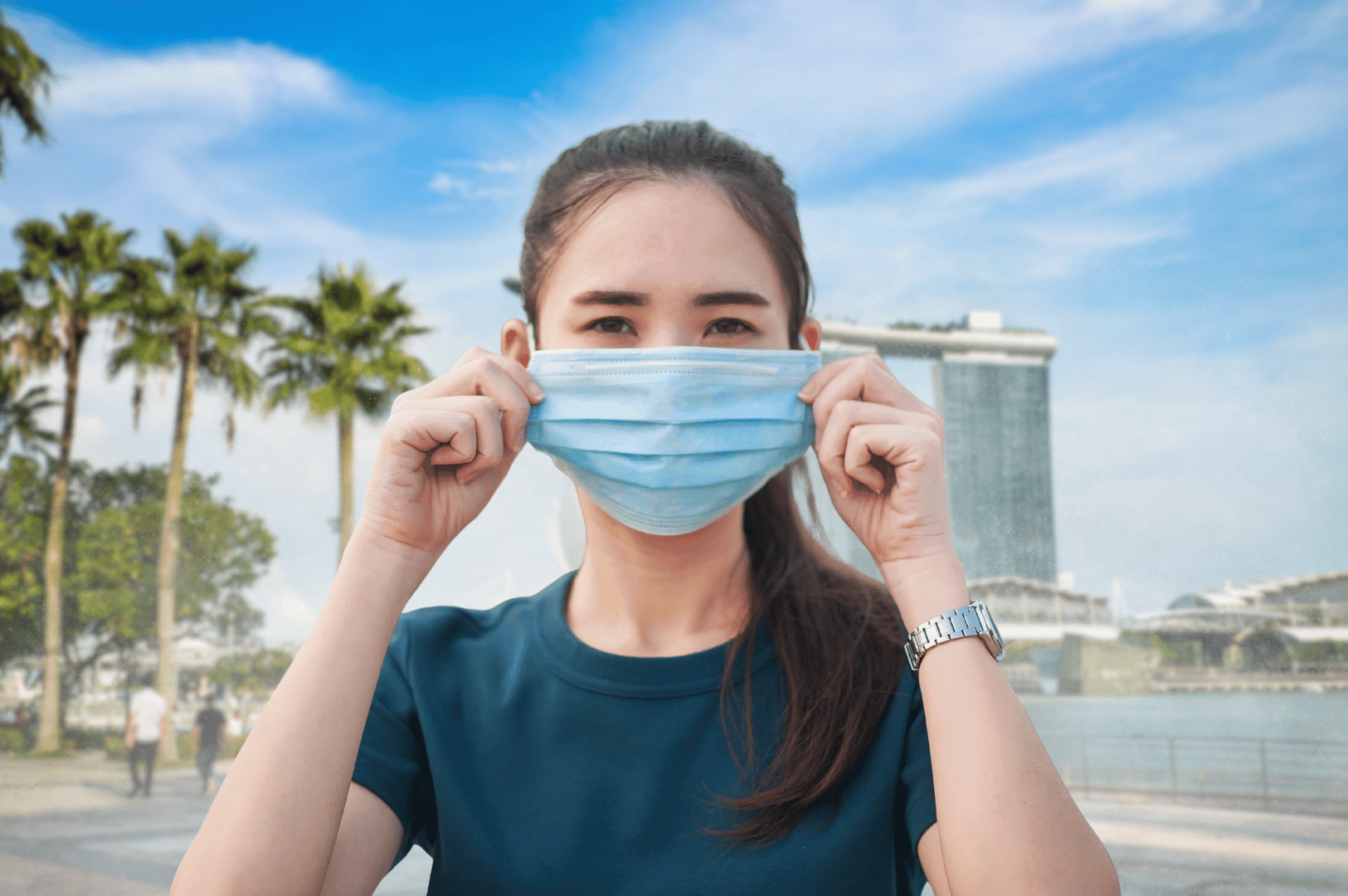 Person outdoors adjusting a face mask on a hazy day, with city skyline and palm trees in the background.