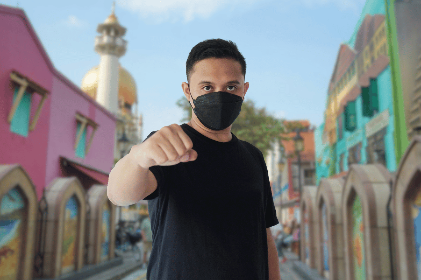 Person wearing a black face mask outdoors, holding a fist forward in a protective gesture, with colorful street buildings in the background.