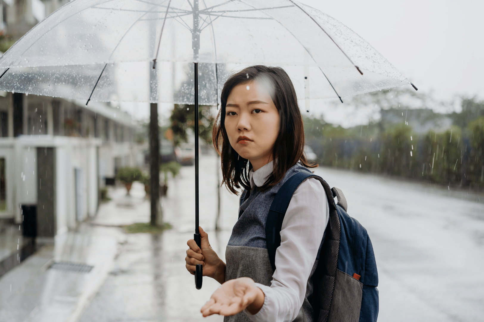 Young commuter under a clear umbrella during a heavy Singapore rain, checking raindrops on her hand—fits article on first monsoon tips.