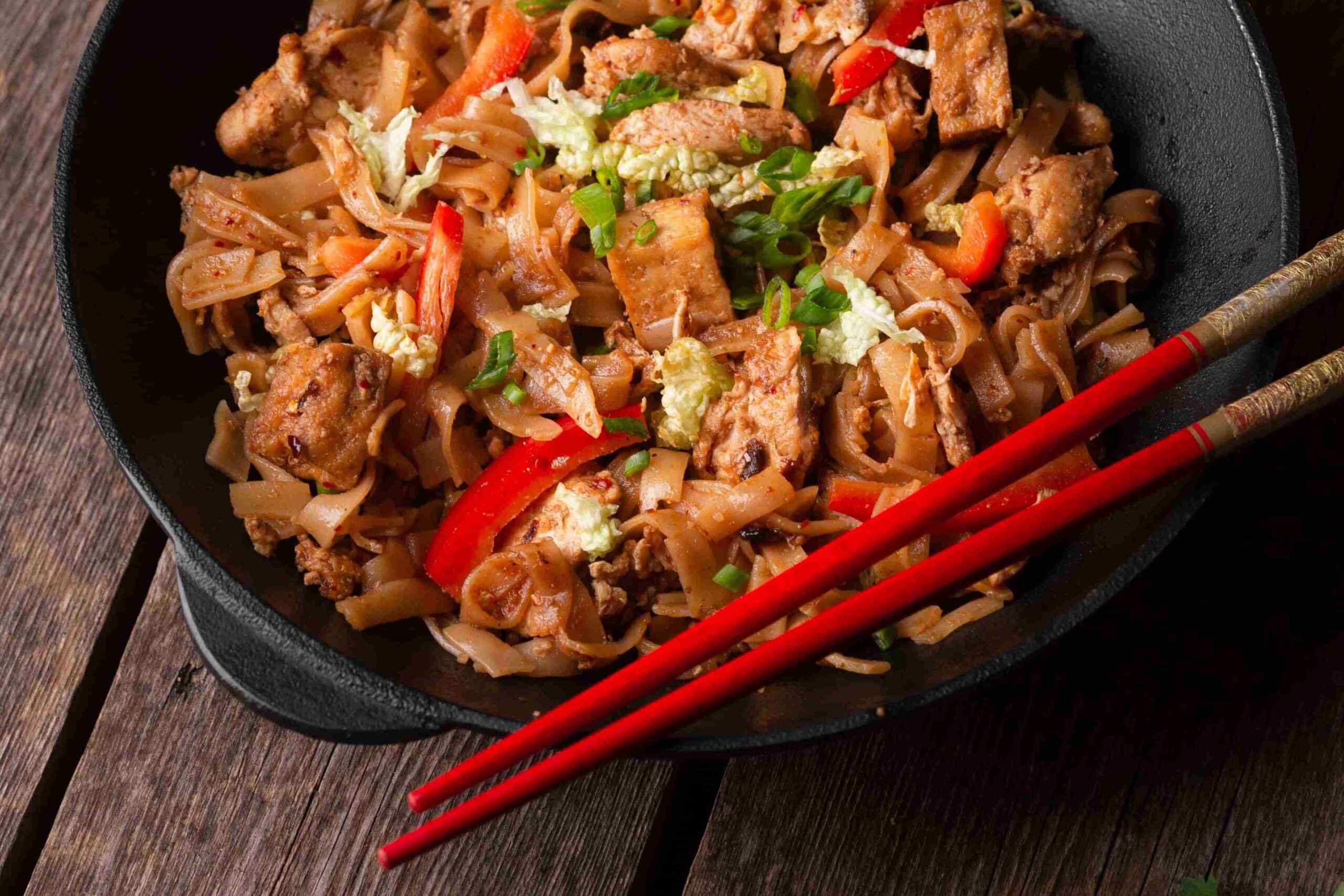 A close-up photo of Char kway teow on black vintage pot with a pair of red chopsticks.
