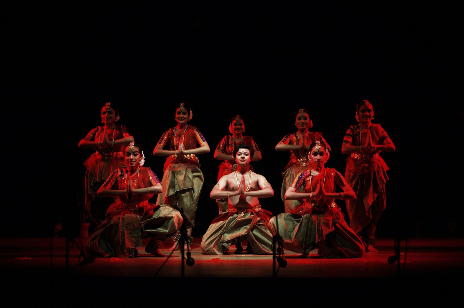 Indian classical dance performance in Singapore with eight dancers in ornate costumes and jewelry, striking symmetrical poses under red stage lighting to showcase the city’s cultural heritage.