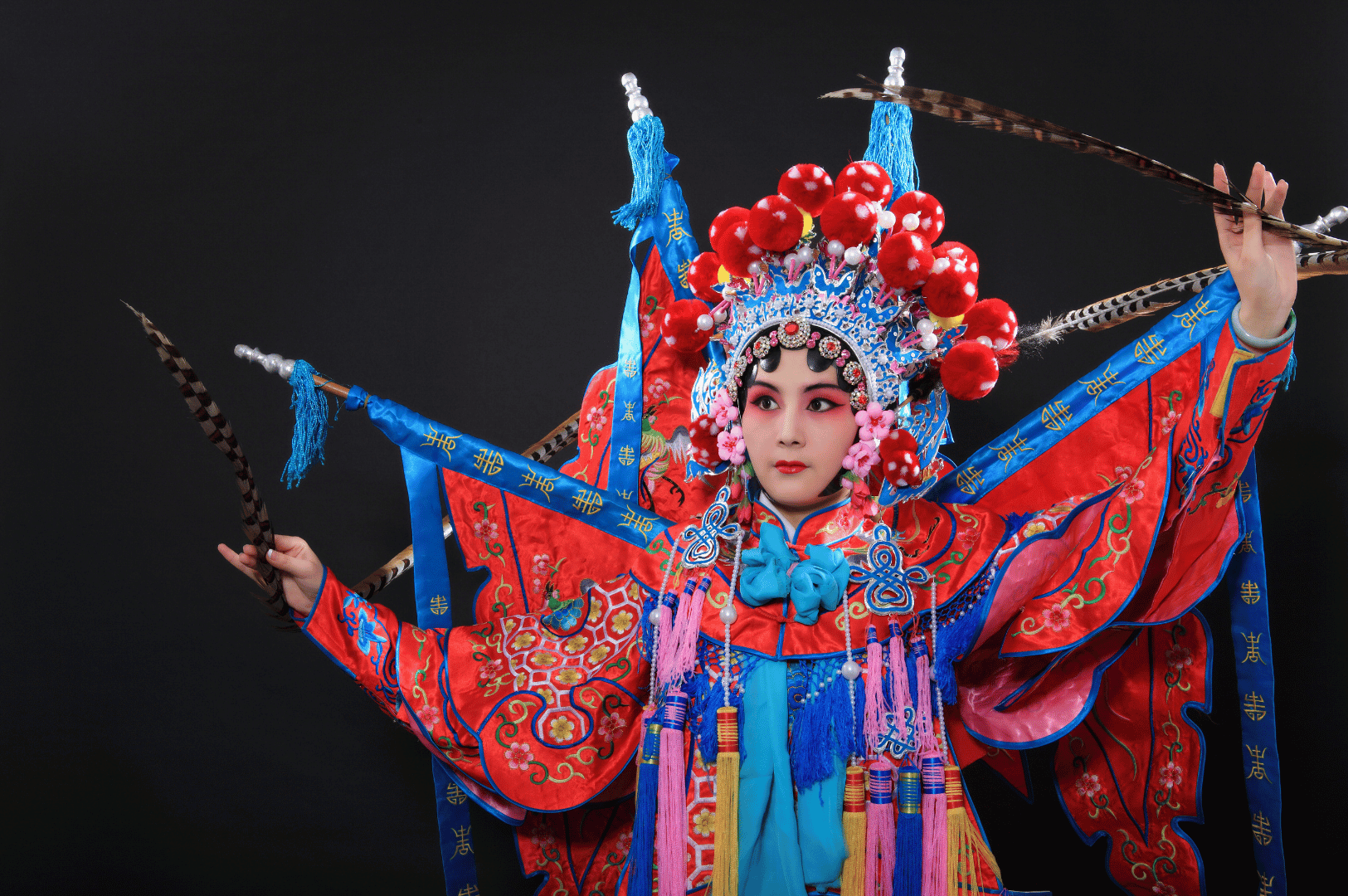 Chinese opera performer in Singapore wearing a vibrant embroidered costume and ornate headdress with red pom-poms, holding long pheasant feathers under stage lighting, representing the city’s rich traditional performing arts.