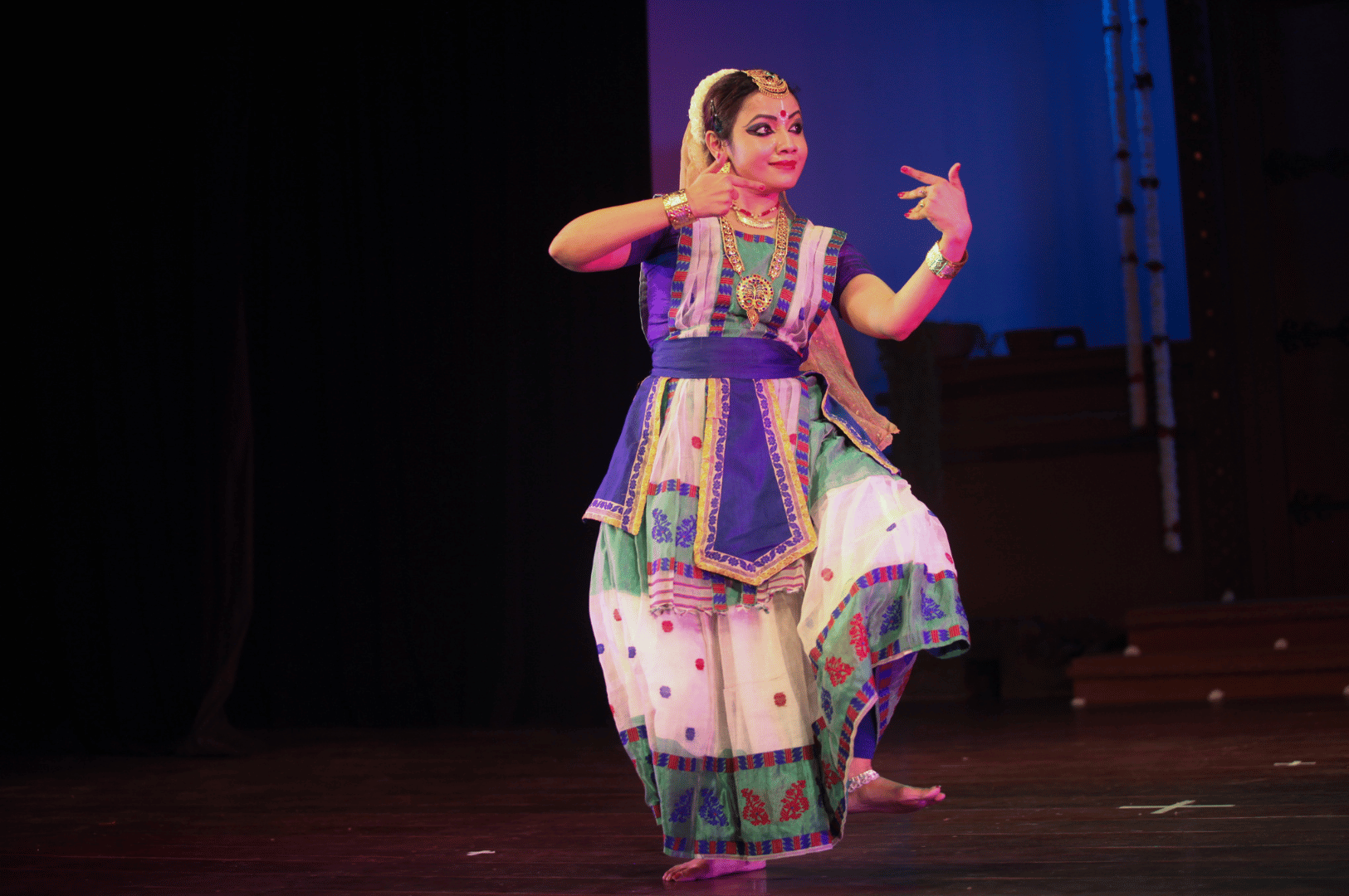 Indian classical dance performance in Singapore featuring a solo dancer in colorful costume with gold embellishments, expressive hand gestures, and dynamic posture under stage lighting, highlighting the city’s cultural heritage.