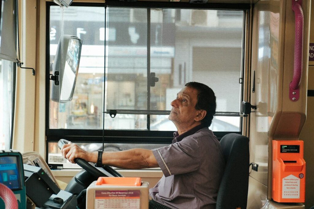 A male bus driver operating a Singapore bus, with a cityscape visible through the windshield.