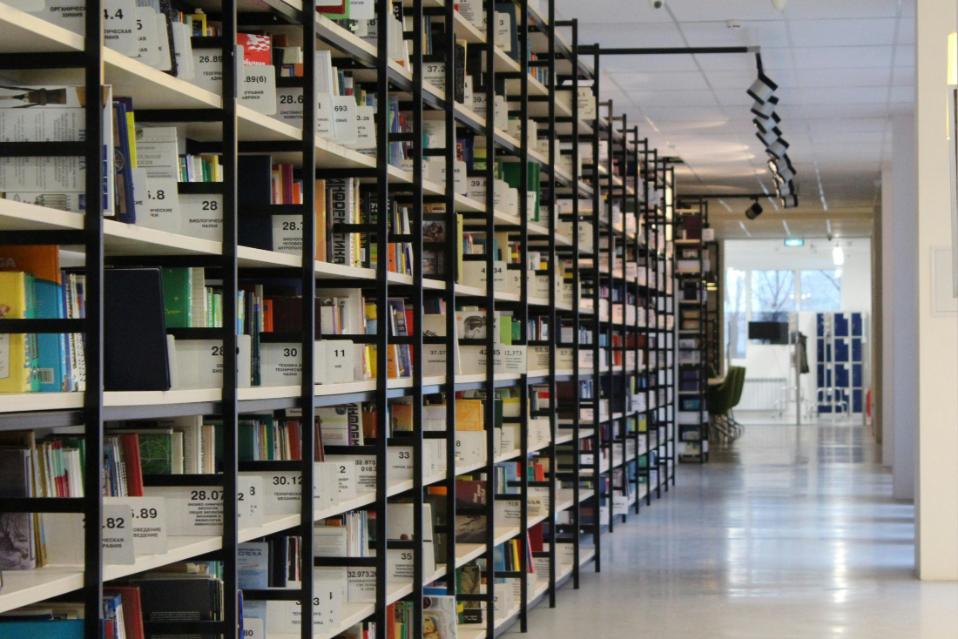 Tall, black metal shelves filled with organized books line the left side of a modern library aisle, stretching deep into the room. To the right, a polished floor reflects the overhead track lighting, leading the eye toward a bright seating area in the distance.