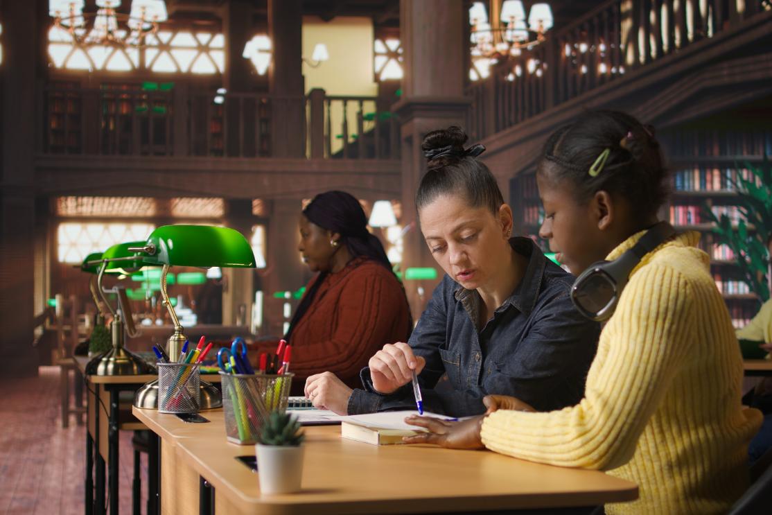 A female tutor assists an elementary school student at a library desk, pointing at a notebook with a blue pen to explain a lesson.