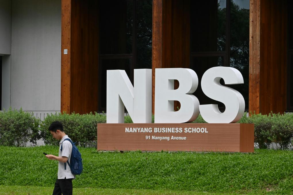 A student on his phone walking past the large sign of the school's name in Singapore.