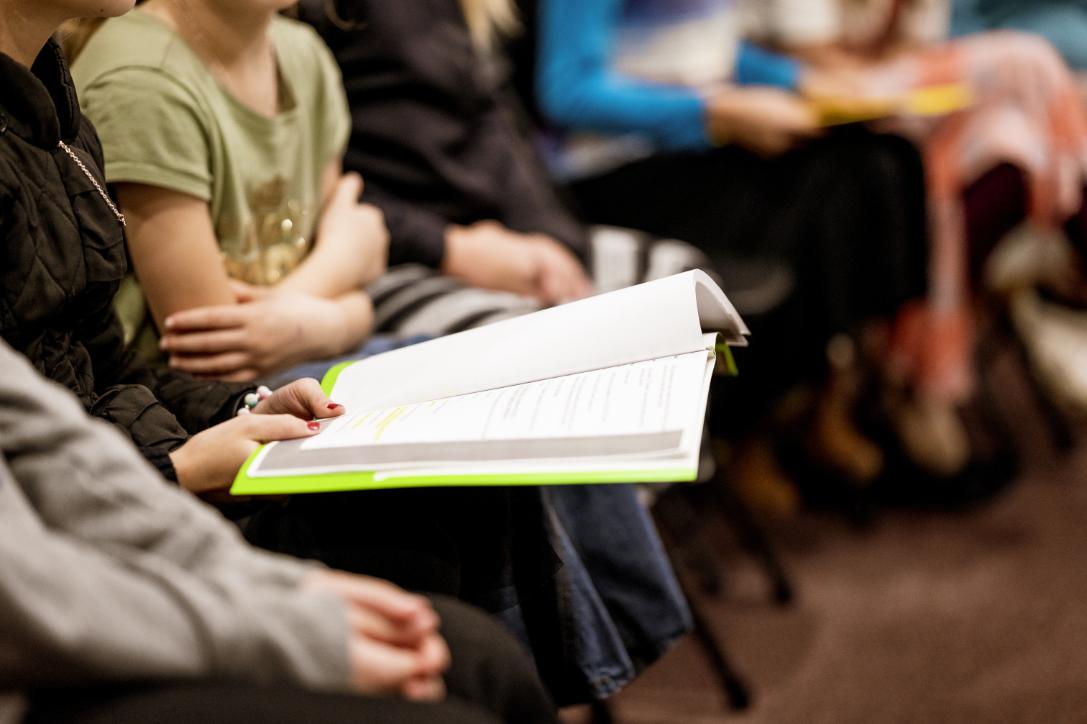 This selective focus shot highlights the hands of a seated individual holding an open booklet or folder with a bright green spine.