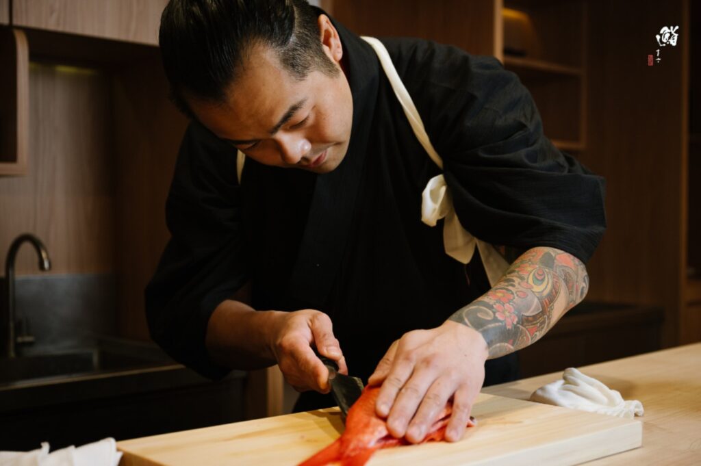 Chef Masa, adorned with tattoos, expertly slices sushi, highlighting his skill and dedication to the art of sushi making.