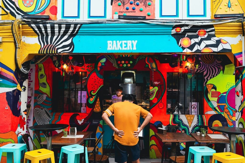 A man stands in front of a vibrant bakery on Haji Lane, Singapore, showcasing colorful pastries and decor.
