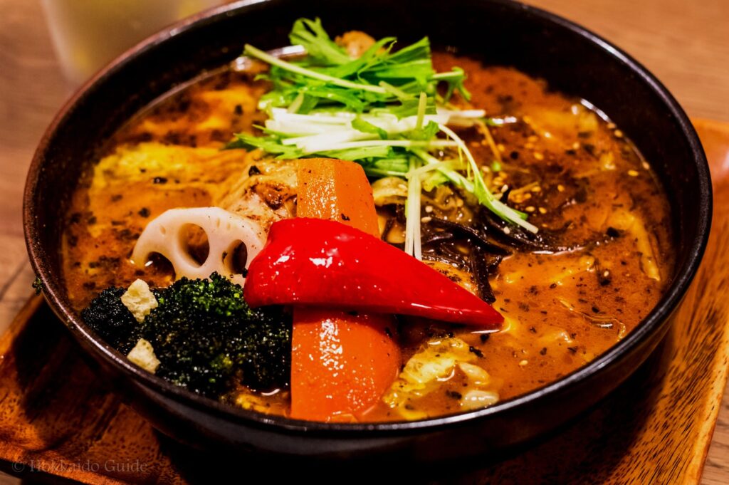 A steaming bowl of curry soup with vibrant vegetables, including red pepper, carrot, lotus root, broccoli, and greens. The dish appears flavorful and inviting.