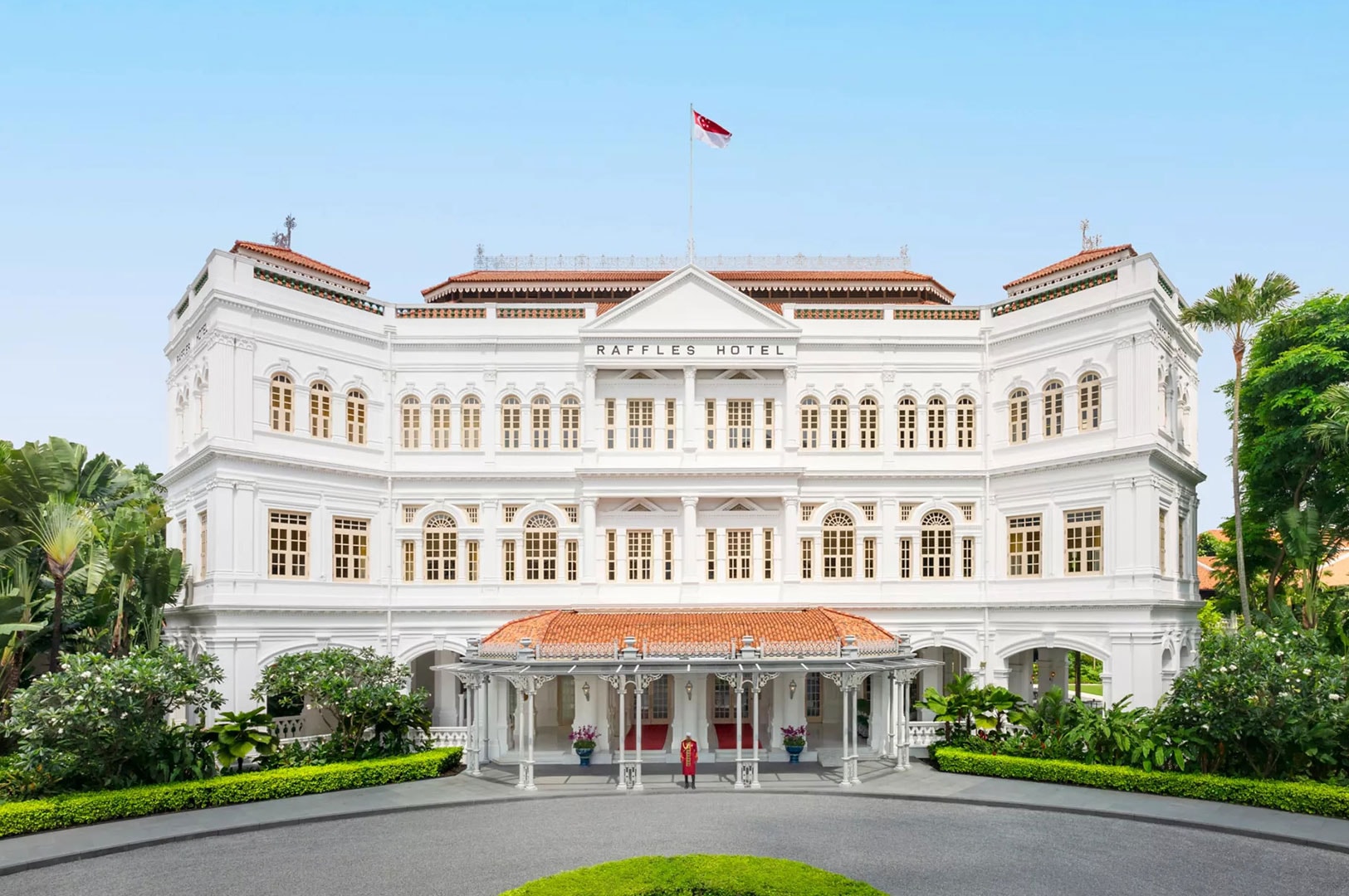 Front view of Raffles Hotel Singapore, a white colonial-style building with arched windows, red-tiled roof, and landscaped entrance under the Singapore flag.