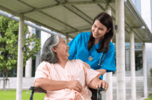 Smiling healthcare professional in blue scrubs stands beside an older adult in a wheelchair under a shaded walkway, symbolizing compassionate elder care within Singapore’s healthcare system. Trees and modern architecture in the background reflect the city’s integration of nature and infrastructure.